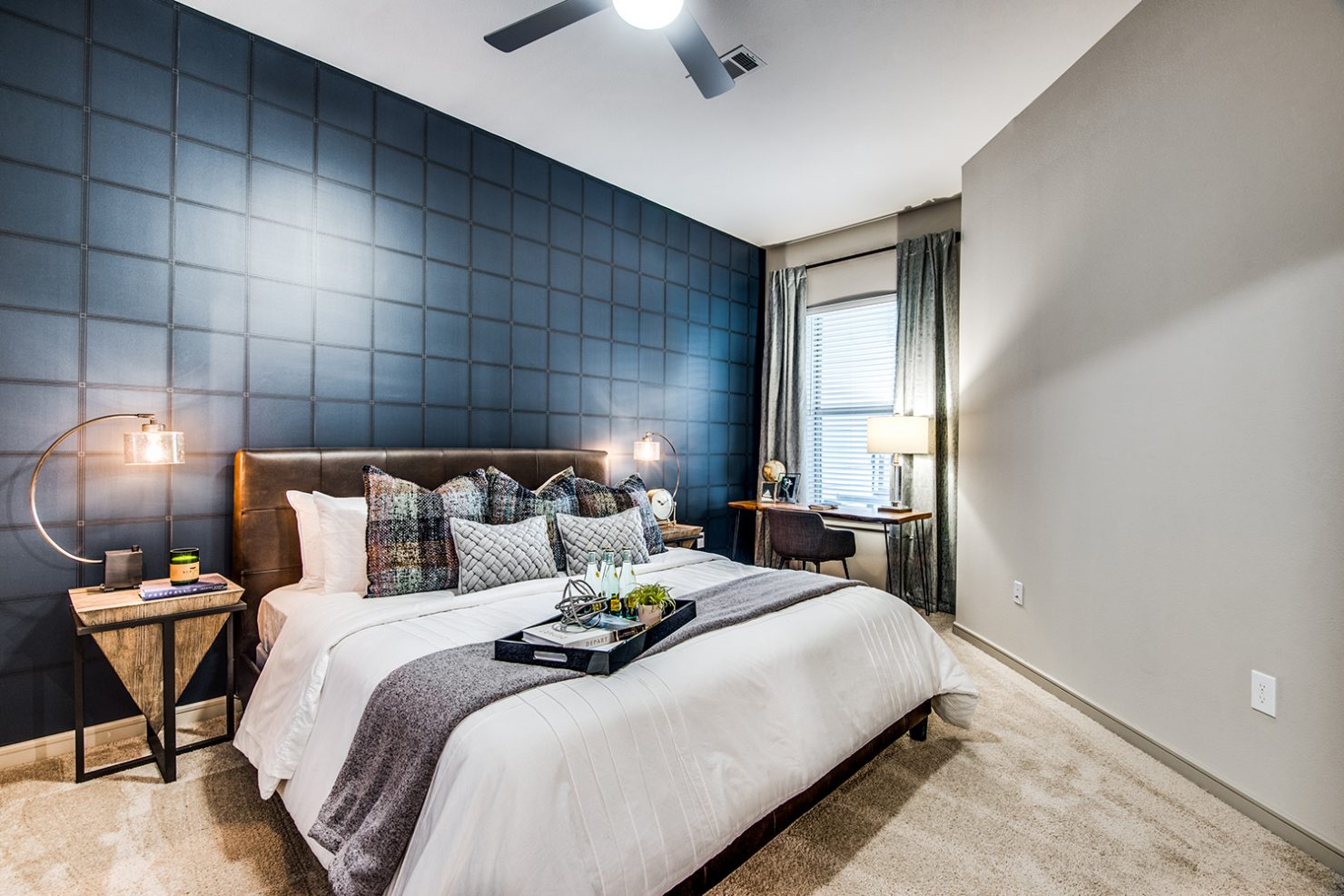 Model apartment bedroom with queen bed, work desk, ceiling fan, and carpeted floor