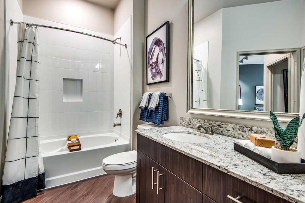 Model apartment bathroom with wood-style floors, granite countertops, brushed nickel hardware, large framed mirror and ceramic tile tub