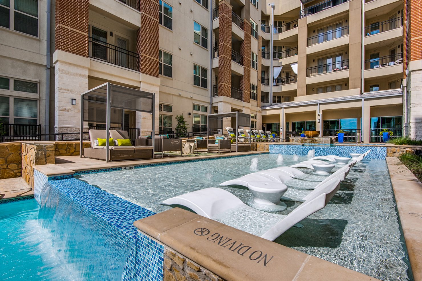 Resort style pool with covered deck seating, waterfall, and in-pool loungers with view of apartments above