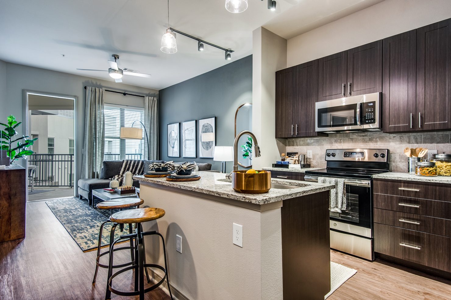 Model apartment kitchen and living area with large island, stainless steel appliances, wood-style flooring, and access to patio or balcony