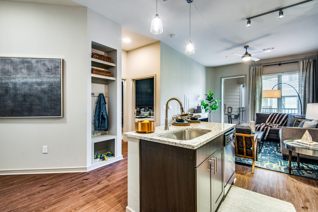 Model apartment with open kitchen with island, living area with designer pendant lighting, wood-style floors, and built-in shelving with coat hooks