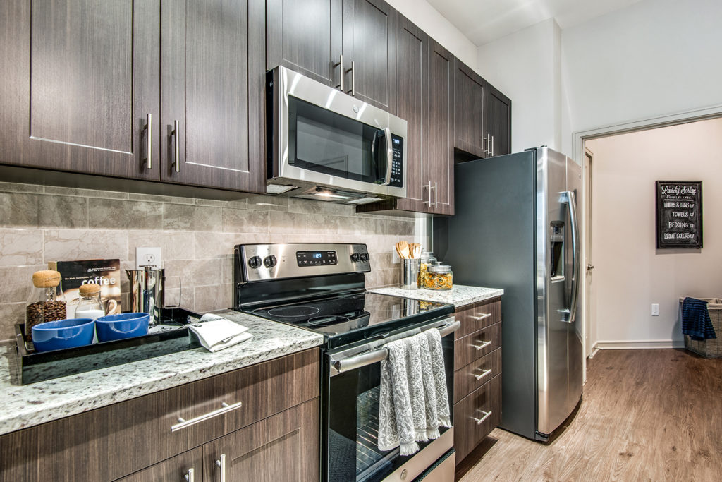 Model apartment kitchen with dark wood cabinetry, designer backsplash, and stainless steel appliances with large refrigerator