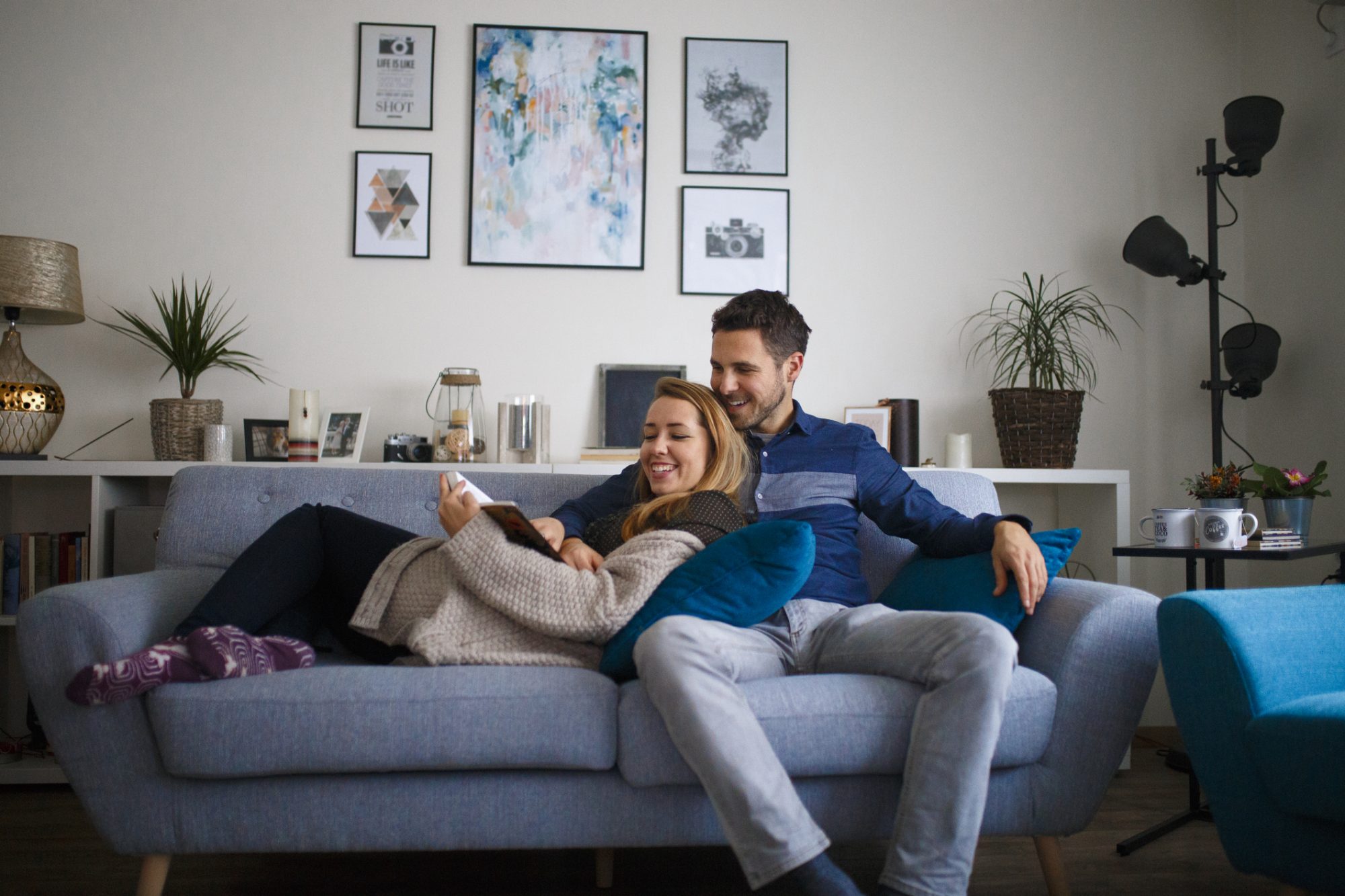 Young couple sitting together on their living room sofa at home reading a book